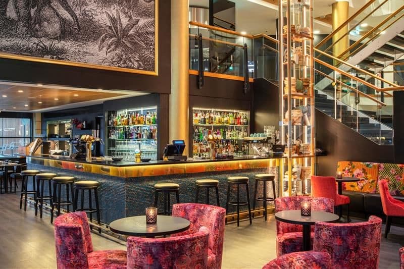 A bar with bar stools, small round tables with red chairs, and a staircase in the background